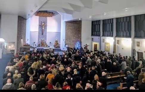 an image of a large number of the congregation sitting on brown church pews during holy Mass